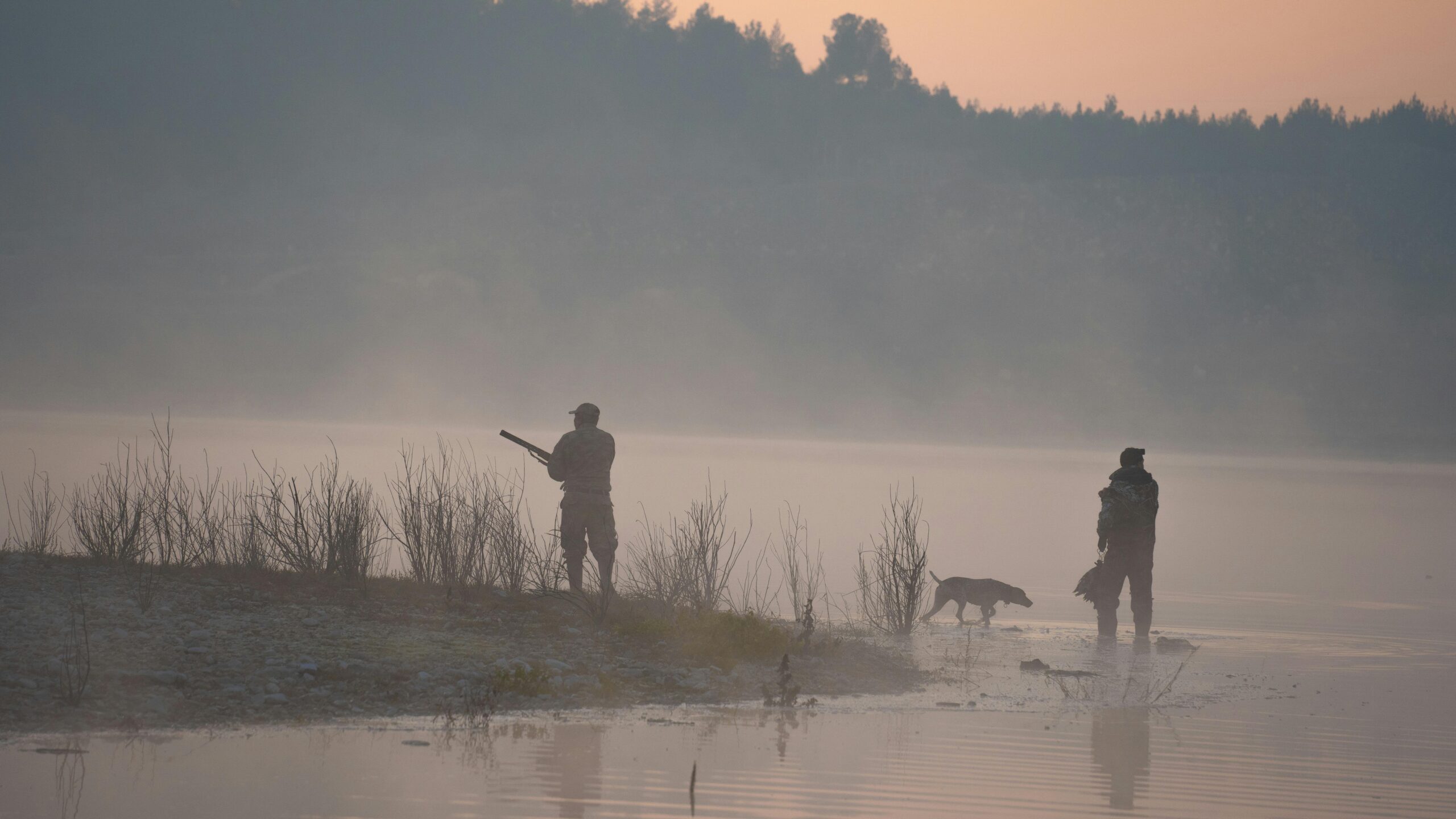 Hunters with a dog by a foggy lakeshore at sunrise, capturing a serene atmosphere.